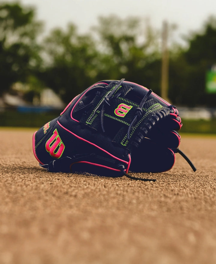 The Wilson A2000 DP15SS 11.5" Baseball Glove (SPRING 2025: WBW102747115) with green and pink accents rests on the infield dirt, featuring Pedroia Fit Technology, with blurred trees and field equipment in the background.