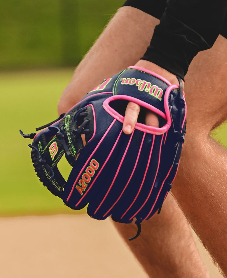 A close-up of a person wearing a Wilson A2000 DP15SS 11.5" Baseball Glove (SPRING 2025: WBW102747115), holding it open with their fingers. The blurred background features green grass on an outdoor field.