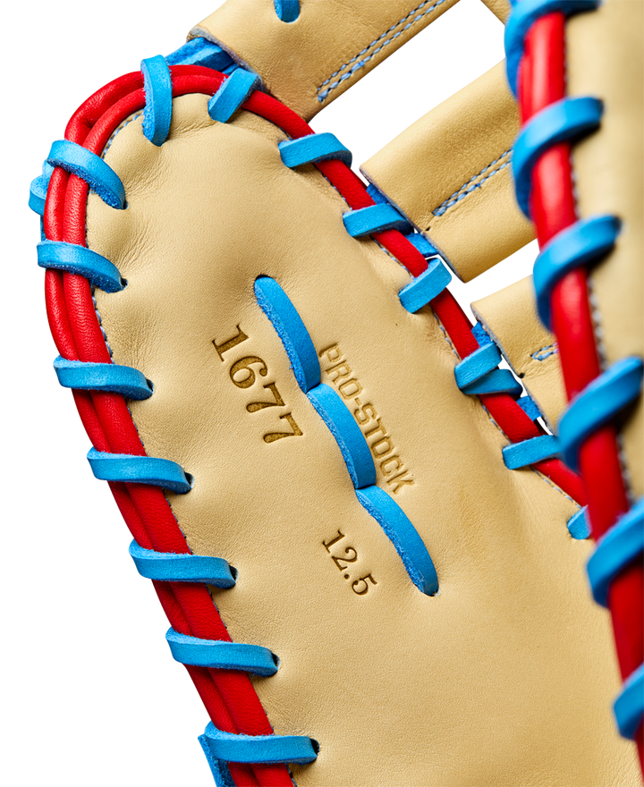 Close-up of the Wilson A2000 1677 12.5" First Base Mitt, featuring tan Pro Stock leather with blue and red lacing, and "1677," "PRO STOCK," and "12.5" stamped in gold on the palm. Model: WBW102753125.