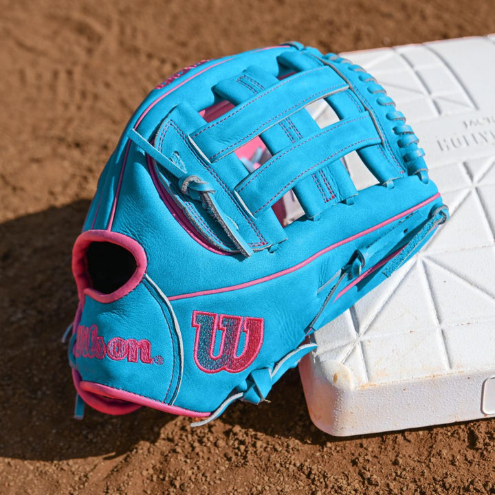 A Wilson A2000 EDLC44 12" Elly De La Cruz Gameday Baseball Glove (FALL 2025: WBW10345212) by Wilson, in blue and pink, rests on a white base on a dirt baseball field.