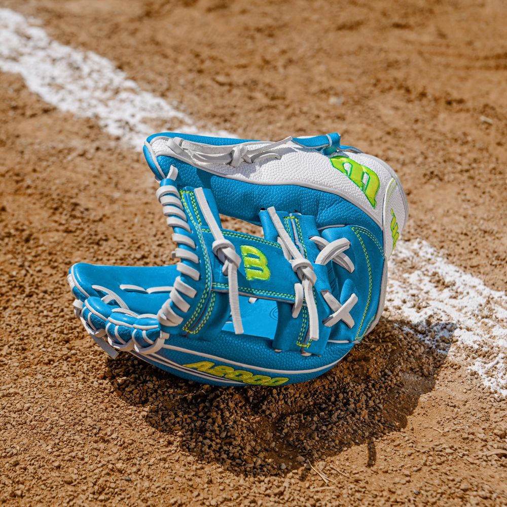 A Wilson A2000 H12SS 12" Fastpitch Glove (FALL 2025: WBW10348612), blue and white with yellow accents, lies open on a dirt field near a white chalk baseline.