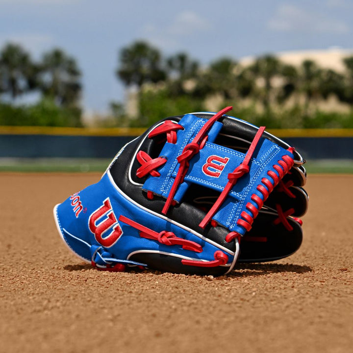 A Wilson A2K MB1175 11.75" Mookie Betts Gameday Baseball Glove rests on the dirt of a baseball field, its blue, black, and red Pro Stock Select leather contrasting with a blurred background of trees and a fence under a clear sky.