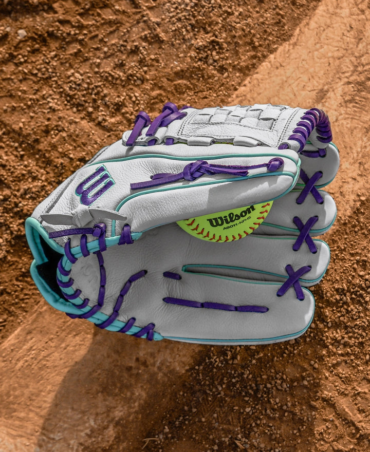 A Wilson A500 Siren 12" Fastpitch Glove: WBW10355912, made of top grain leather with purple and teal laces, holds a yellow softball on a dirt infield near the baseline, ready for action.