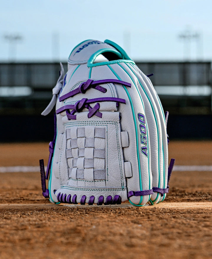 The Wilson A500 Siren 12" Fastpitch Glove (WBW10355912) is a white youth glove with teal and purple accents, crafted from top grain leather, standing upright on a dirt field with a blurred stadium fence and lights behind it.