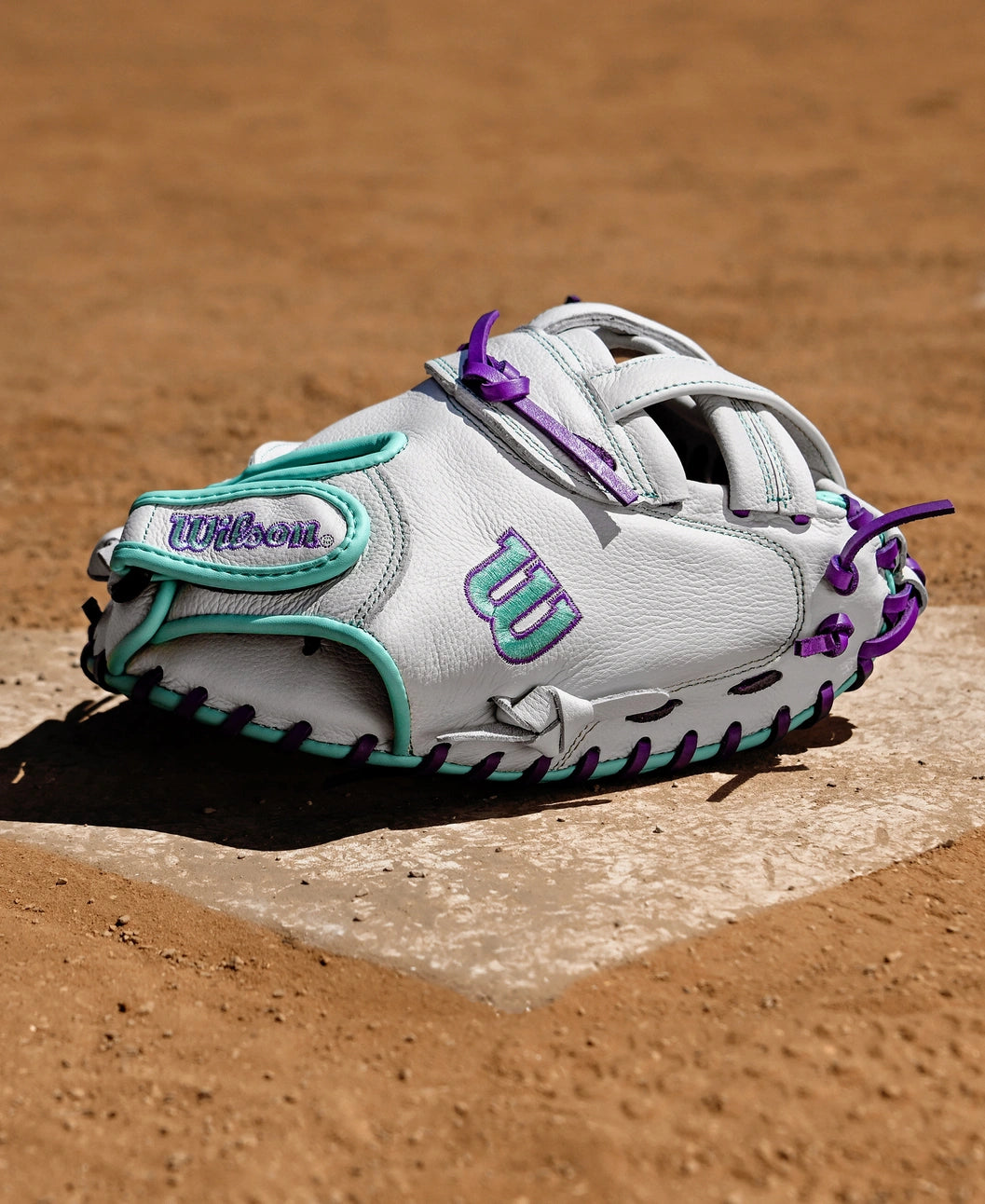 A Wilson A500 Siren 32" Fastpitch Catcher's Mitt (WBW10356332), white with teal and purple accents, rests on home plate of a dirt field.