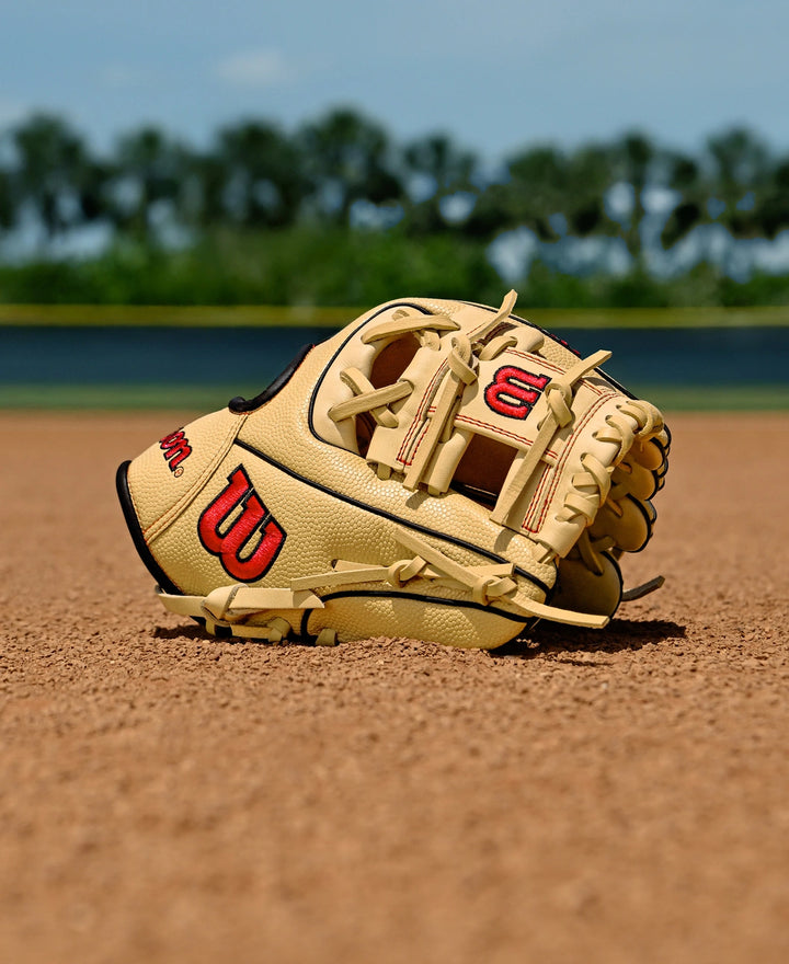 A Wilson A1000 PF11 11" Baseball Glove by Wilson, tan with red and black accents, rests on a baseball field’s dirt, surrounded by green grass and trees under a blue sky.