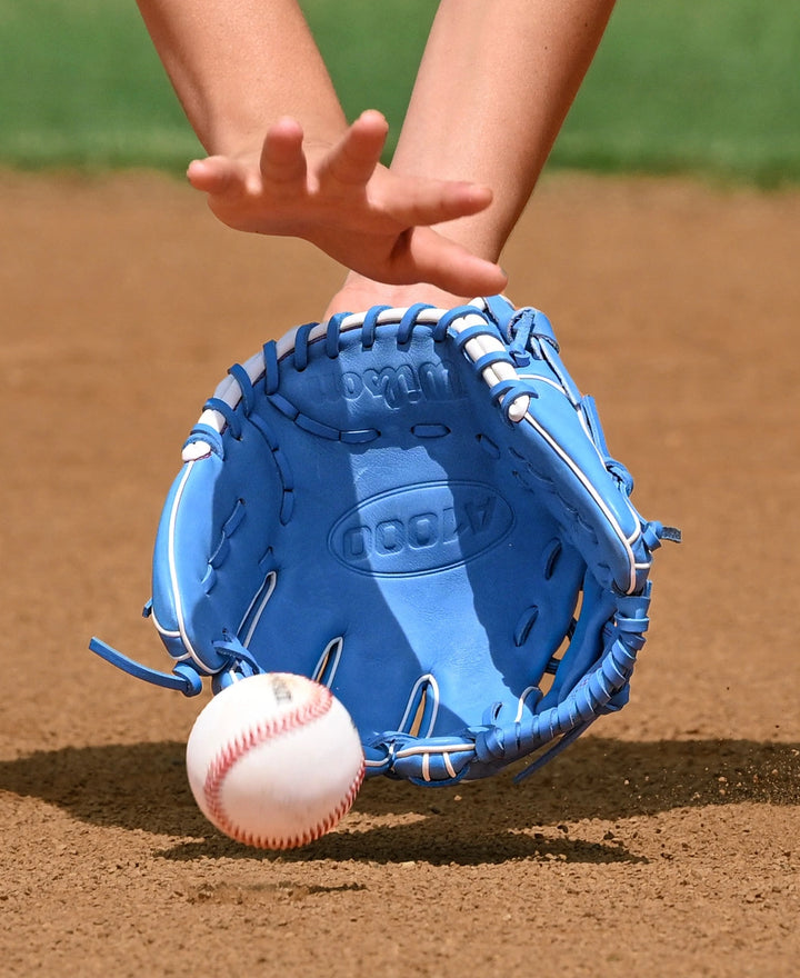Wearing a Wilson A1000 1786 11.5" Baseball Glove (WBW103957115), a player reaches for a bouncing ball on brown dirt, with green grass in the background.