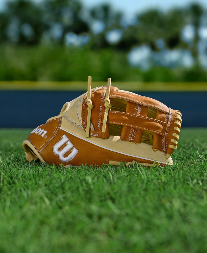 A tan and brown Wilson A1000 1750 12.5" Baseball Glove (WBW103964125) rests on green grass, with a blurred background of trees and a distant fence beneath a bright sky.