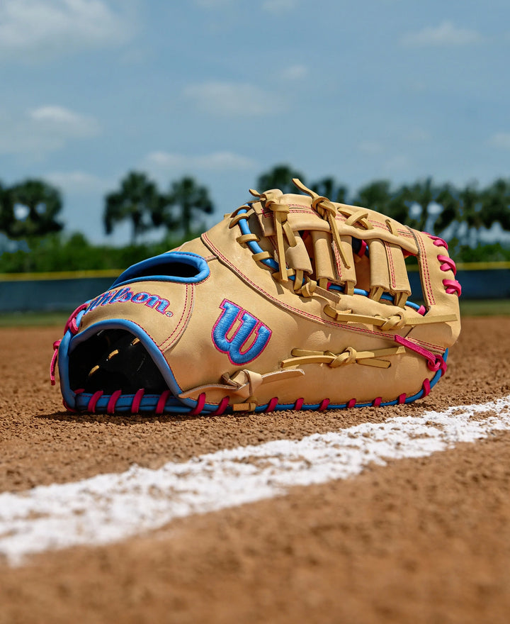 A Wilson A1000 1620 12.5" First Base Mitt by Wilson, in tan and blue full grain leather with pink laces, lies on the dirt near a white foul line on a baseball field, with green trees and a blue sky in the background.