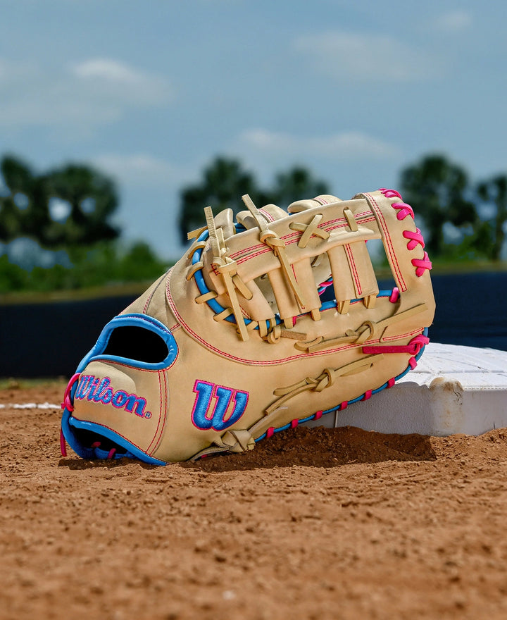 A Wilson A1000 1620 12.5" Baseball First Base Mitt by Wilson, in beige with blue and pink laces, sits on a base on a dirt field, framed by green trees and a blue sky in the background.