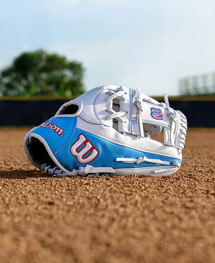 A Wilson A1000 H12 12" Fastpitch Glove (WBW10396912) by Wilson in white and blue rests on the infield dirt of a baseball field, with blurred trees and a fence visible in the background.