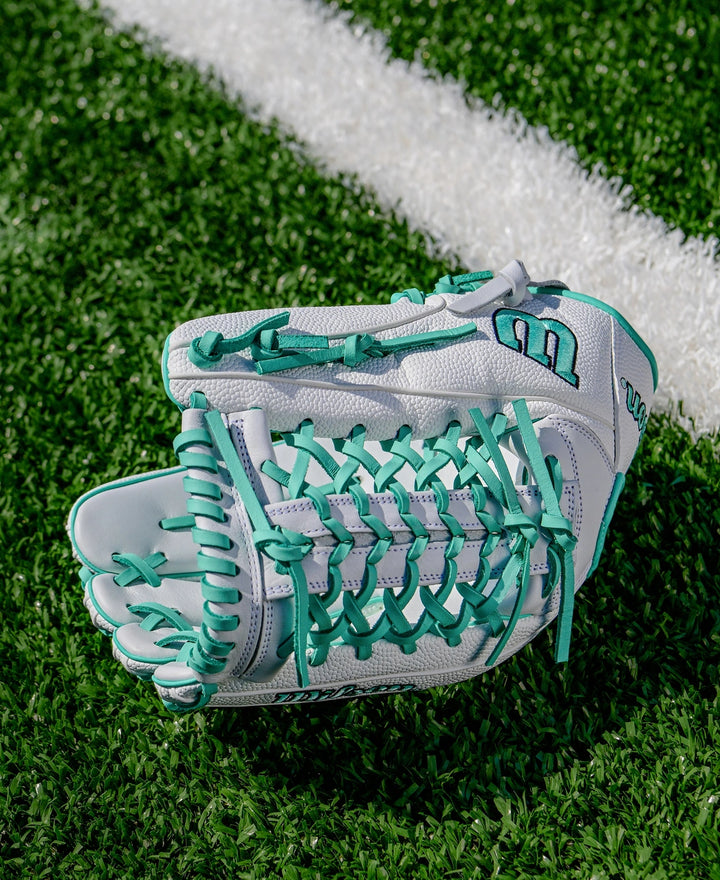 A Wilson A1000 T125 12.5" Fastpitch Glove (WBW103970125), featuring white full grain leather and teal laces, sits on green artificial turf near a white line.