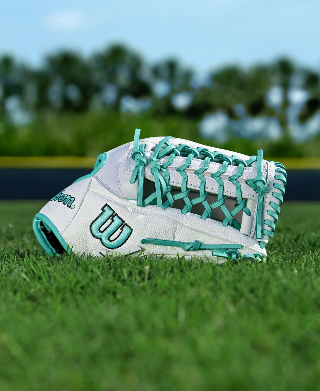 The Wilson A1000 T125 12.5" Fastpitch Glove by Wilson, in white and teal full grain leather, rests on green grass with a blurred background of trees and a dark fence.