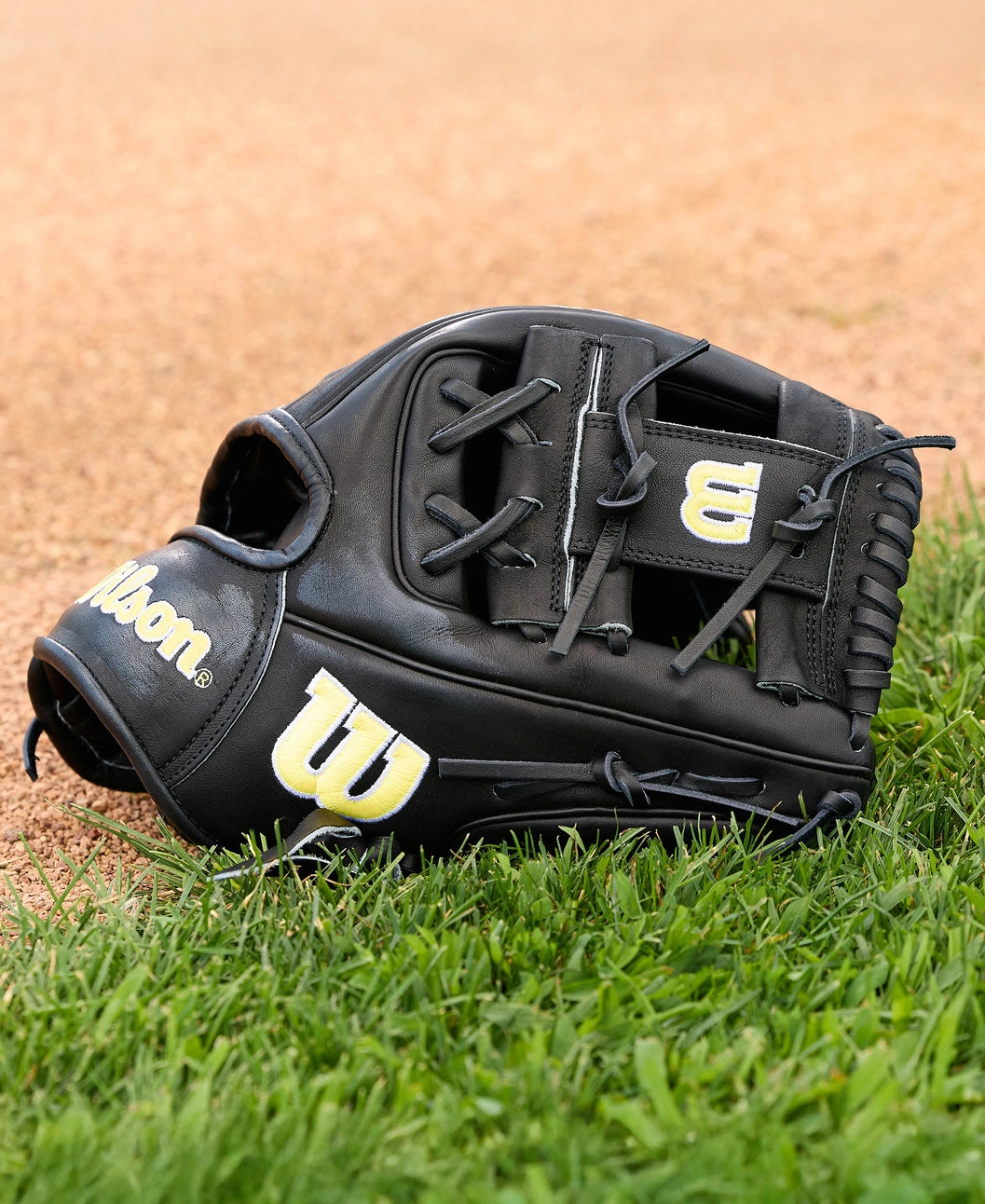 A Wilson A2000 1788 11.25" baseball glove by Wilson, featuring a black design with yellow and white logos, rests on green grass near the dirt infield. Crafted from premium Pro Stock Leather.