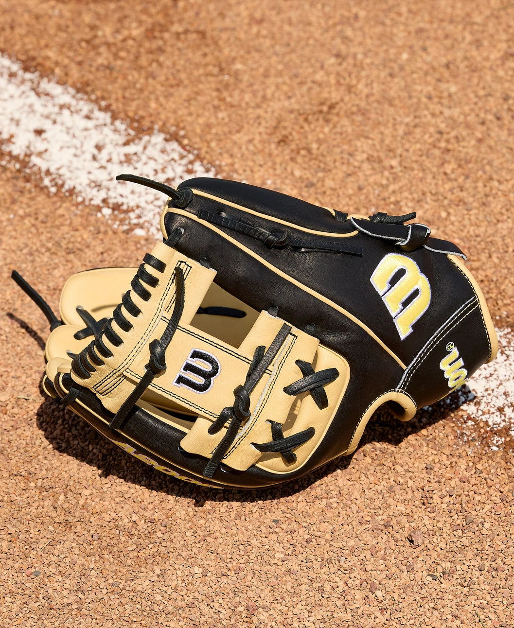 A Wilson A2000 1786 11.5" Baseball Glove (WBW104108115) with black and tan leather and yellow and white Wilson logo rests on reddish-brown dirt beside a white chalk line on a baseball field.