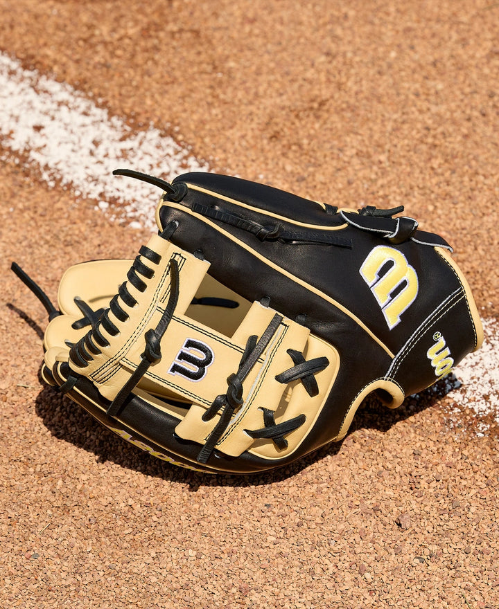 A Wilson A2000 1786 11.5" Baseball Glove (WBW104108115) with black and tan leather and yellow and white Wilson logo rests on reddish-brown dirt beside a white chalk line on a baseball field.