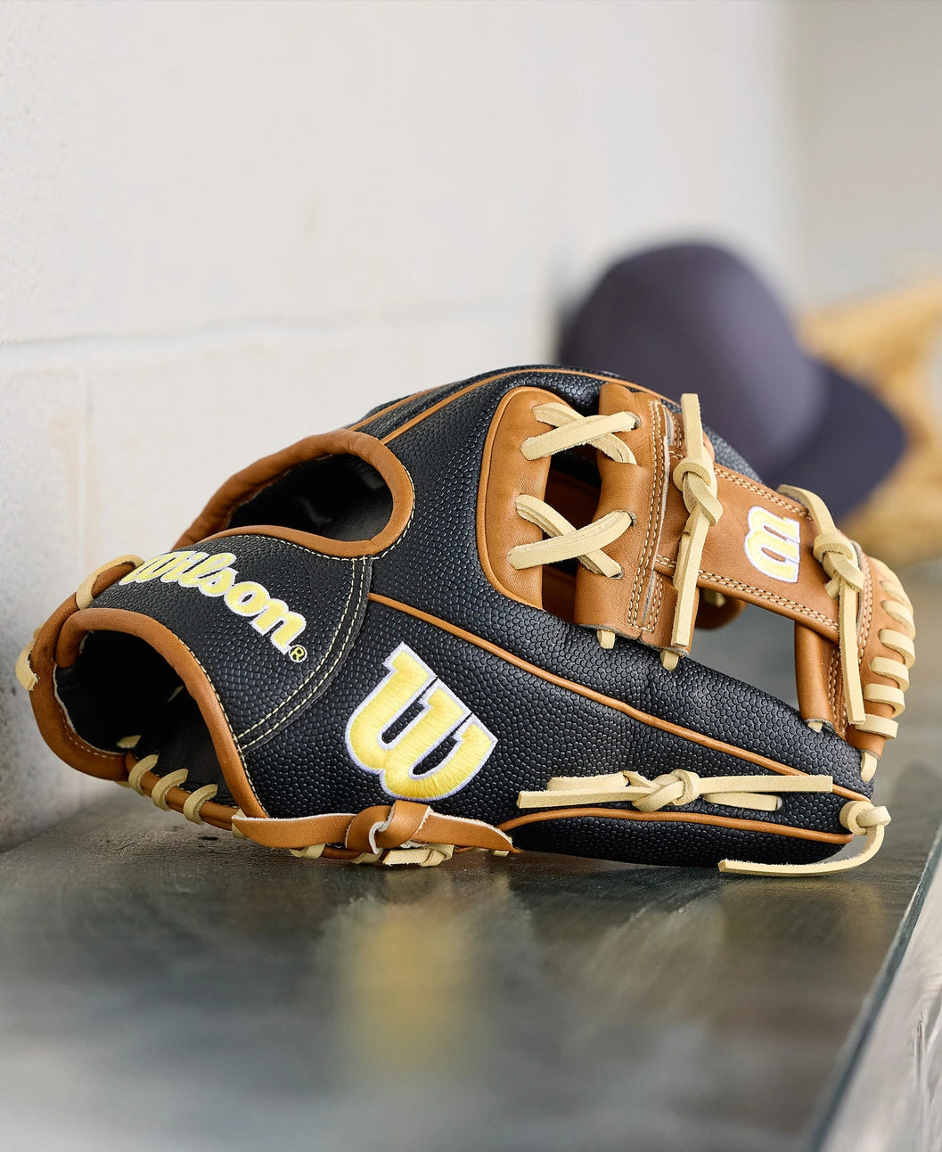 A Wilson A2000 1975SS 11.75" baseball glove (WBW1041121175) by Wilson, featuring black and tan leather with cream laces, sits on a metal dugout surface with a blurred cap visible in the background.