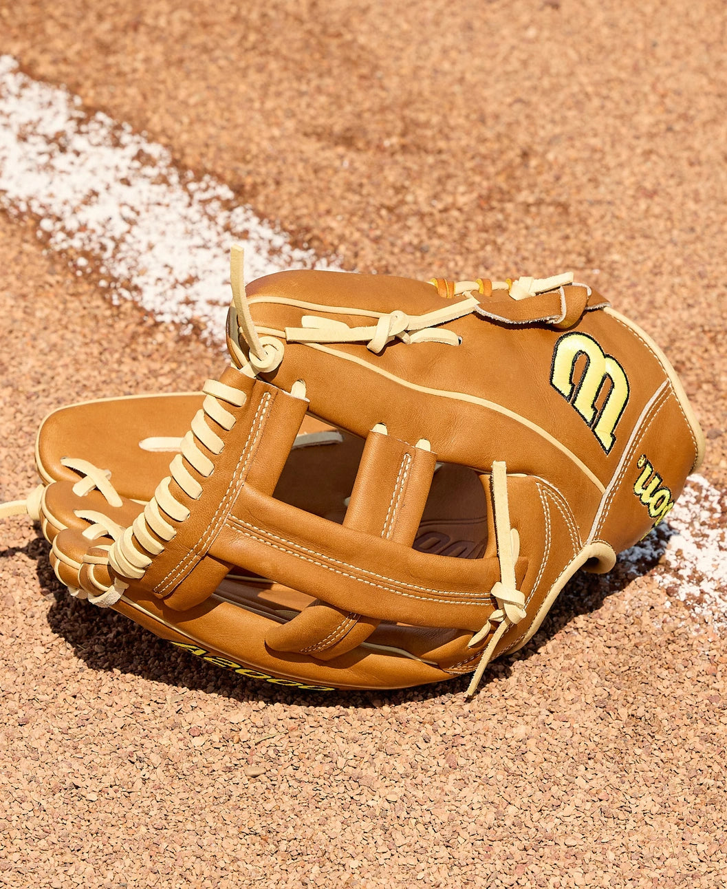 A Wilson A2000 EL3 11.75" baseball glove with tan Pro Stock Leather, white laces, and a yellow W logo rests on the dirt by the chalk baseline.