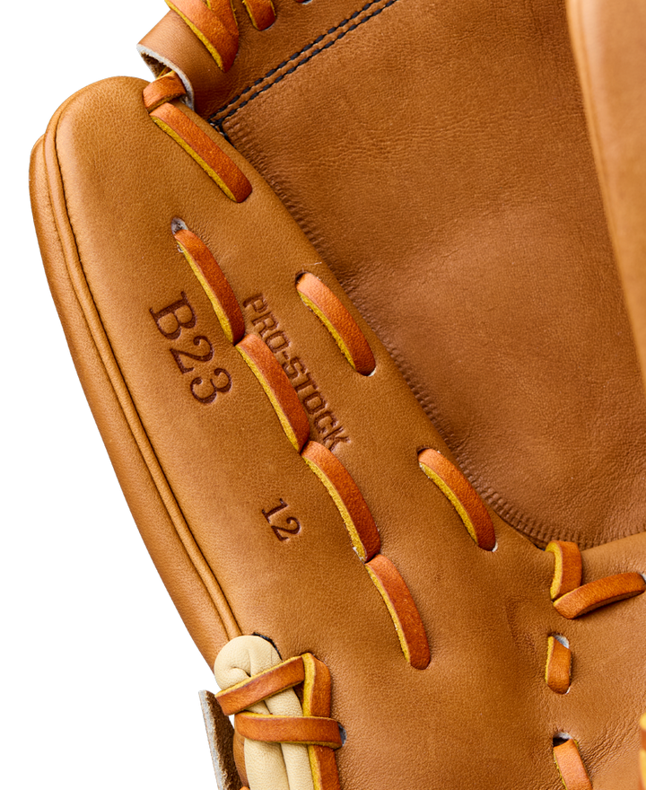 Close-up of a Wilson A2000 B23 12" Baseball Glove with brown Pro Stock leather, tan and orange laces, visible stitching, and embossed text: B23, PRO-STOCK, and 12. Model: WBW10411712.