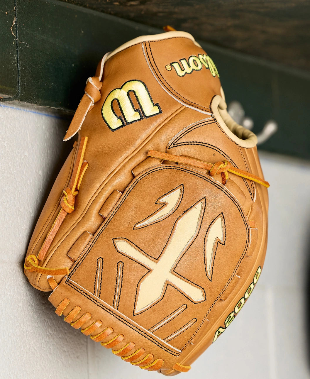 A tan Wilson A2000 B23 12" Baseball Glove (WBW10411712) with yellow stitching and graphic accents hangs on a hook against a white and green wall.