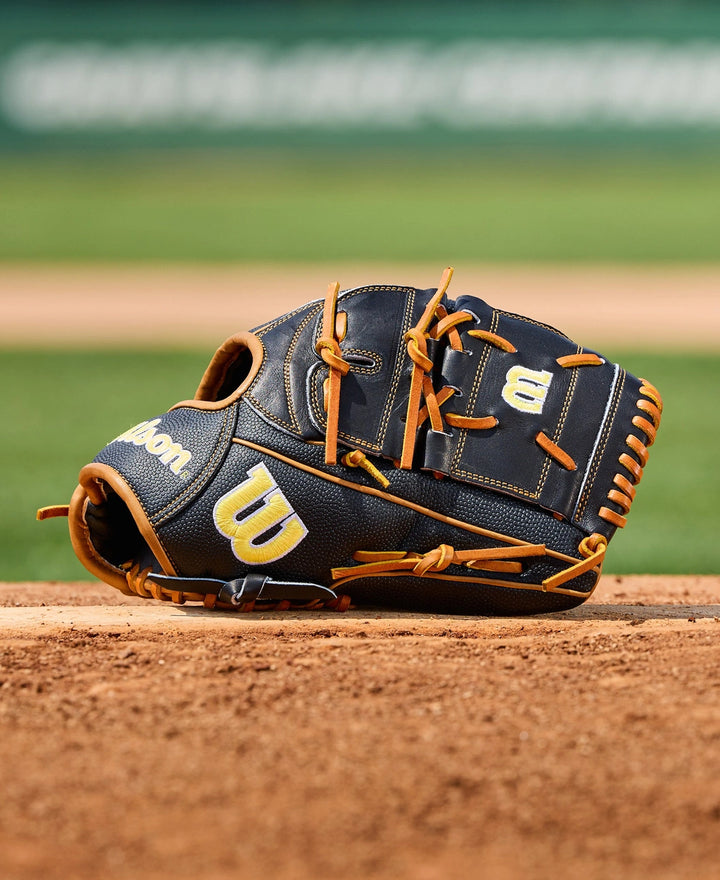 A Wilson A2000 B125SS 12.5" baseball glove (WBW104119125) by Wilson, in black and tan, sits on the dirt of a baseball field with the green outfield blurred in the background.