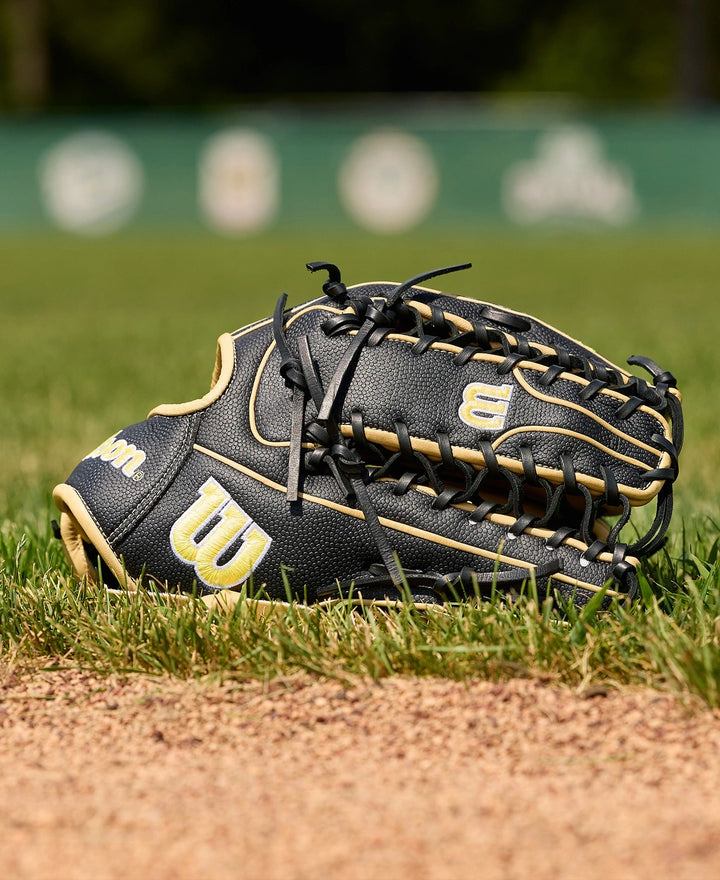 A Wilson A2000 OT7SS 12.75" Baseball Glove (WBW1041831275) by Wilson rests on the grass near the infield dirt, with a blurred green outfield fence and logos in the background.