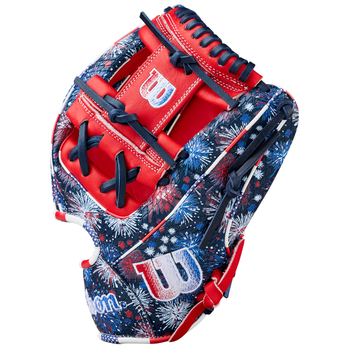 The Wilson A2000 1975SS 11.75" Baseball Glove (GOTM July 2025) features a red, white, and blue fireworks pattern, Pro Stock leather, bold red accents, navy laces, and white W logos on the thumb and wrist.