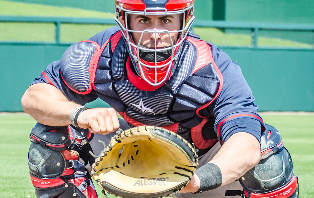 A baseball catcher in full protective gear squats behind home plate, holding a glove open and ready to catch a pitch. The field and green fence are visible in the background.