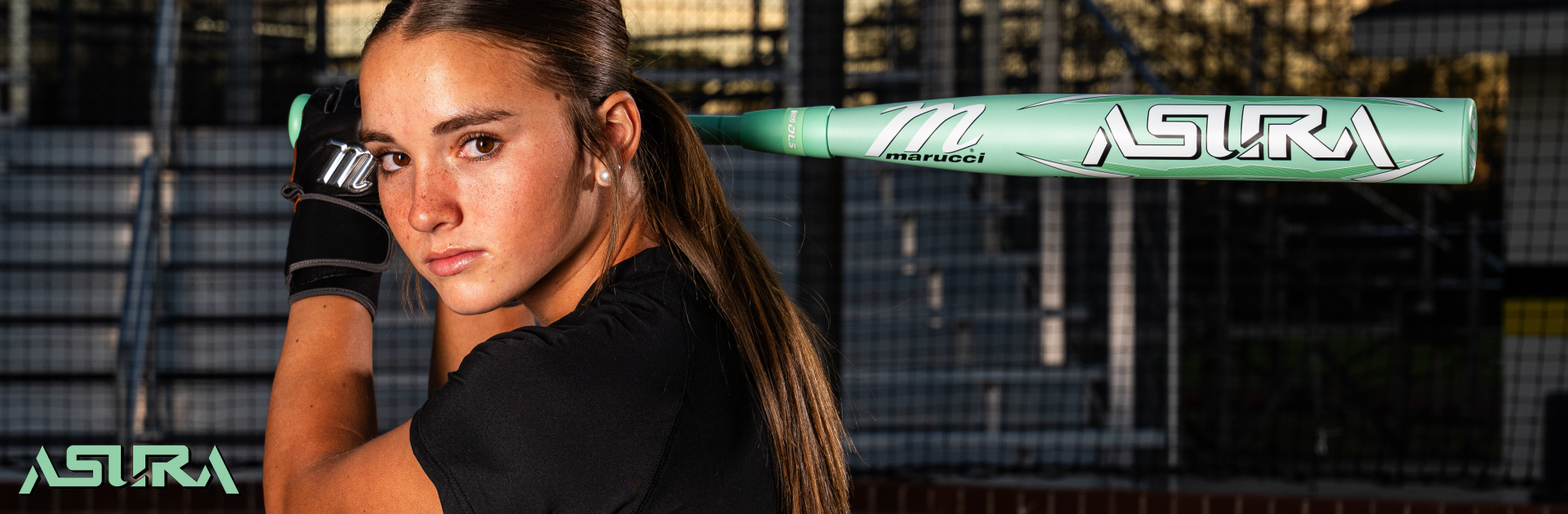 A young woman holding a mint green Marucci ASTRA softball bat over her shoulder, wearing black gloves, looks confidently at the camera with a fenced sports field in the background. The ASTRA logo appears in the corner.