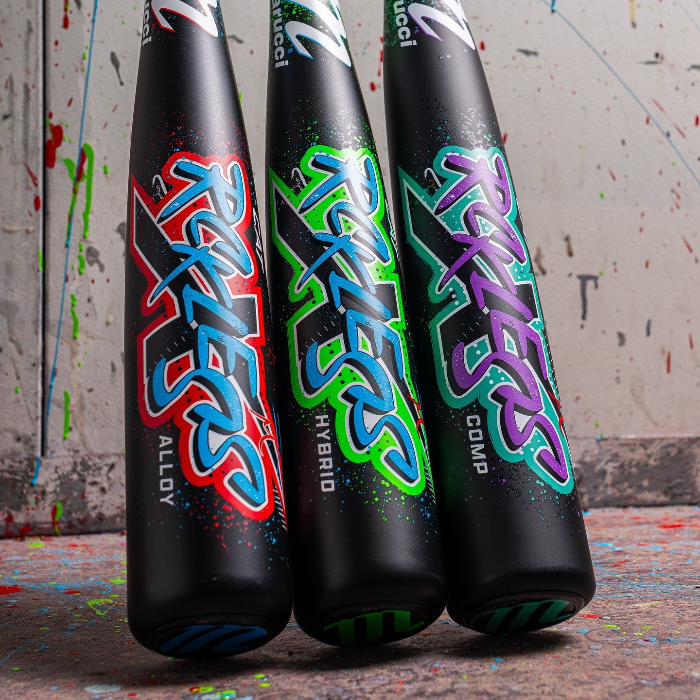 Three black baseball bats with colorful graffiti-style lettering stand upright against a splattered paint background. Each bat is labeled: Alloy (red/blue), Hybrid (green/blue), and Comp (purple/teal).