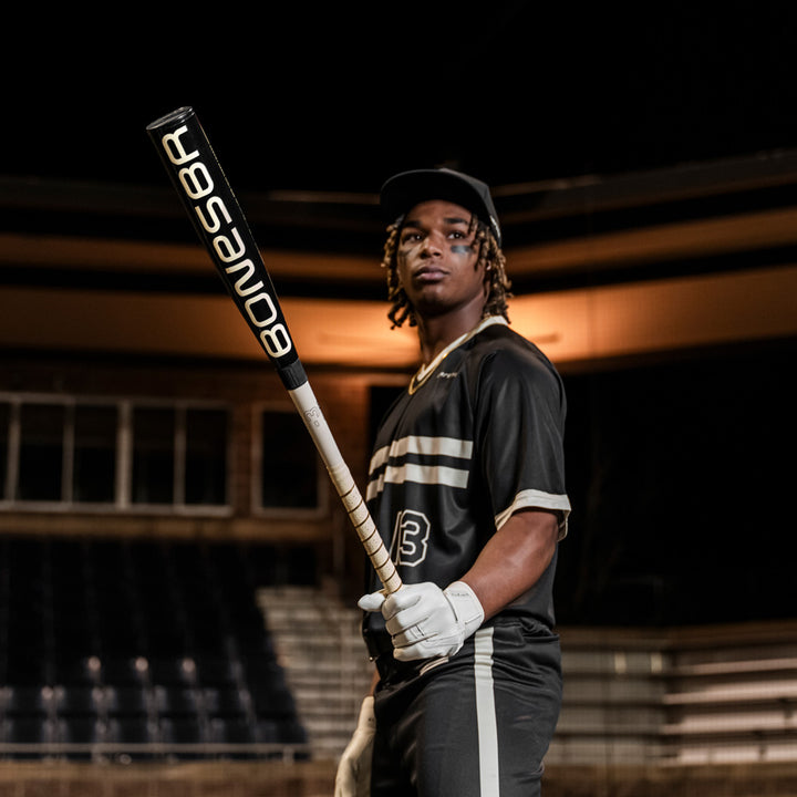 A baseball player in a black uniform with white stripes holds the 2025 Warstic Bonesaber Hybrid Black Cobra (-3) BBCOR Baseball Bat (MBBSRHB25BK3) in a stadium at night, wearing a cap and batting gloves.