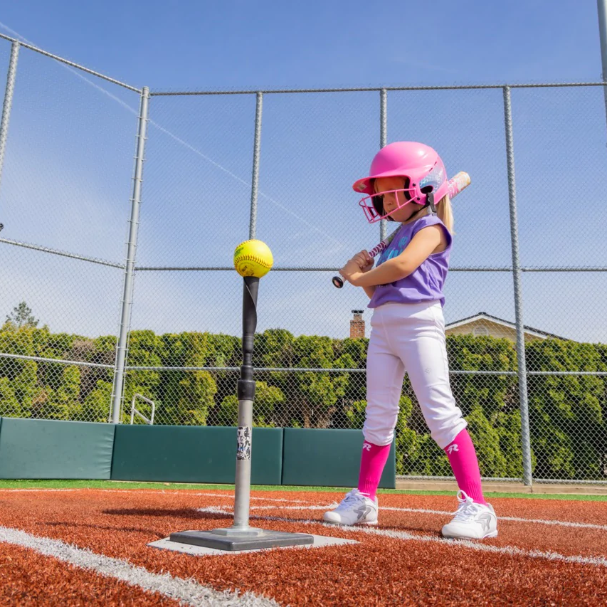 A young child in a pink helmet and purple shirt prepares to hit a softball off a tee with the 2022 Rip It Spark -12 Fastpitch Softball Bat by Rip It at a sunny baseball field.