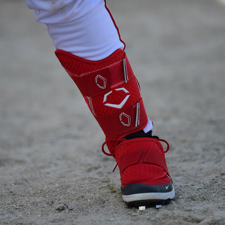 A baseball player's lower leg, dressed in white pants and a red cleat, is shown wearing the EvoShield PRO-SRZ 2.0 Batter's Leg Guard (WB5726901), standing on sandy ground.