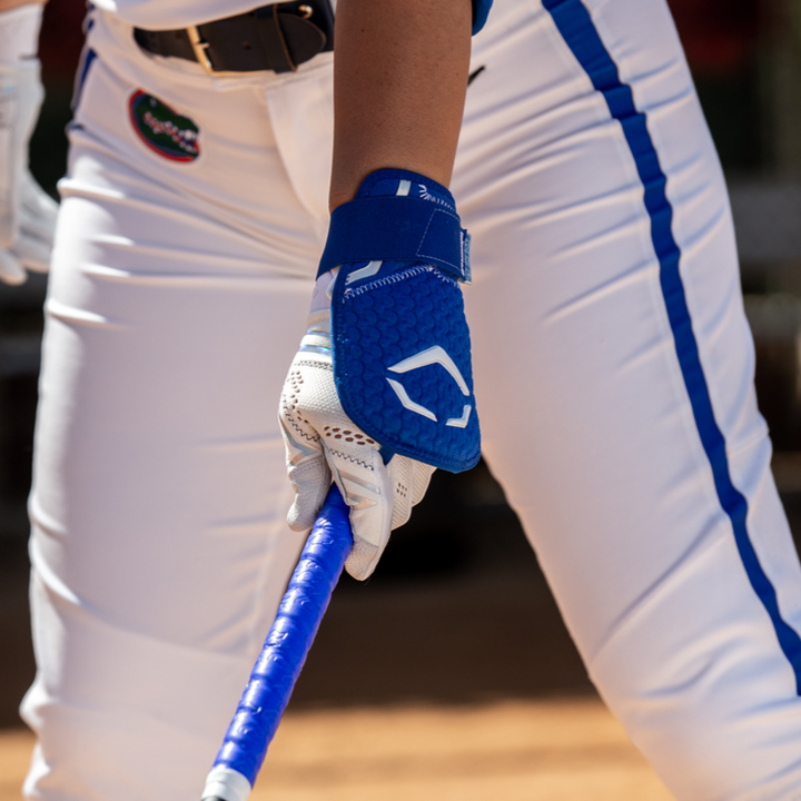 A softball player in white pants with blue stripes grips a bat wrapped in blue tape, wearing the EvoShield PRO-SRZ 2.0 Batter's Hand Guard (WB5726801) for superior protection. The image highlights the lower body and hands.