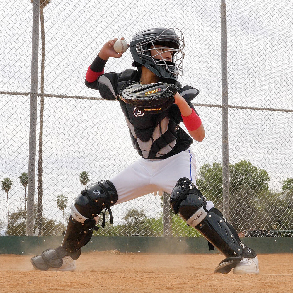 A young baseball catcher in full gear, featuring Force3 Catcher's Leg Guards with Dupont Kevlar (BC8), crouches with a glove, ready to throw a baseball on a dirt field with a chain-link fence and trees behind.