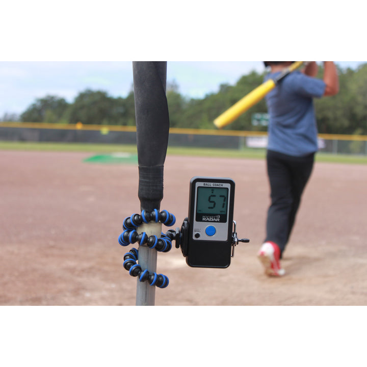 A Pocket Radar Ball Coach Radar (PR1000-BC) by Pocket Radar, showing 57 mph, is mounted on a tripod attached to a bat. In the background, someone swings a yellow bat on a baseball field.