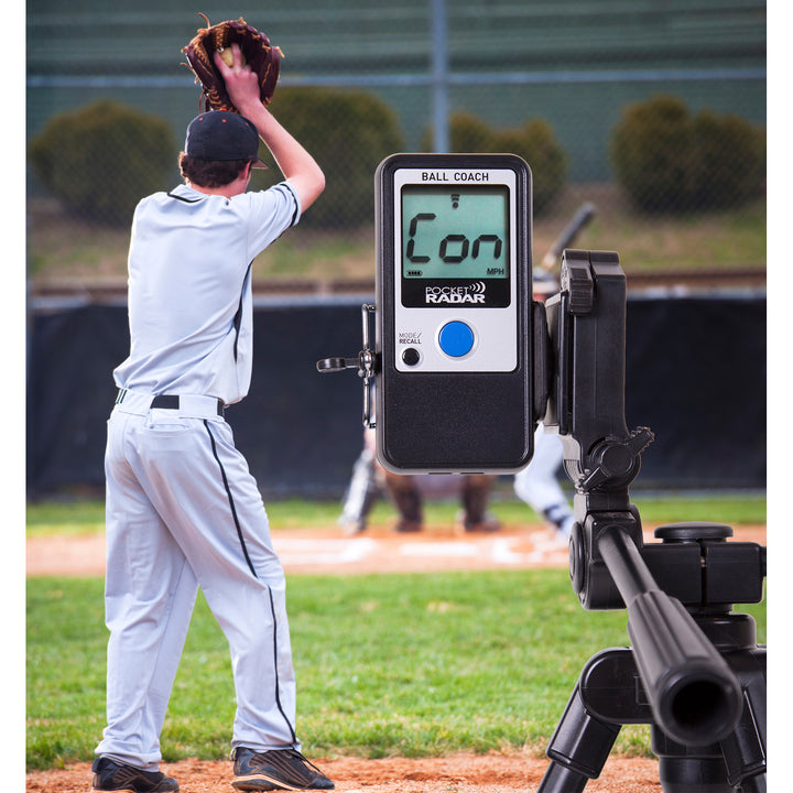 A baseball pitcher winds up on the field while a Pocket Radar Ball Coach Radar (PR1000-BC) by Pocket Radar sits on a tripod in the foreground, its digital screen visible as a key speed training tool.