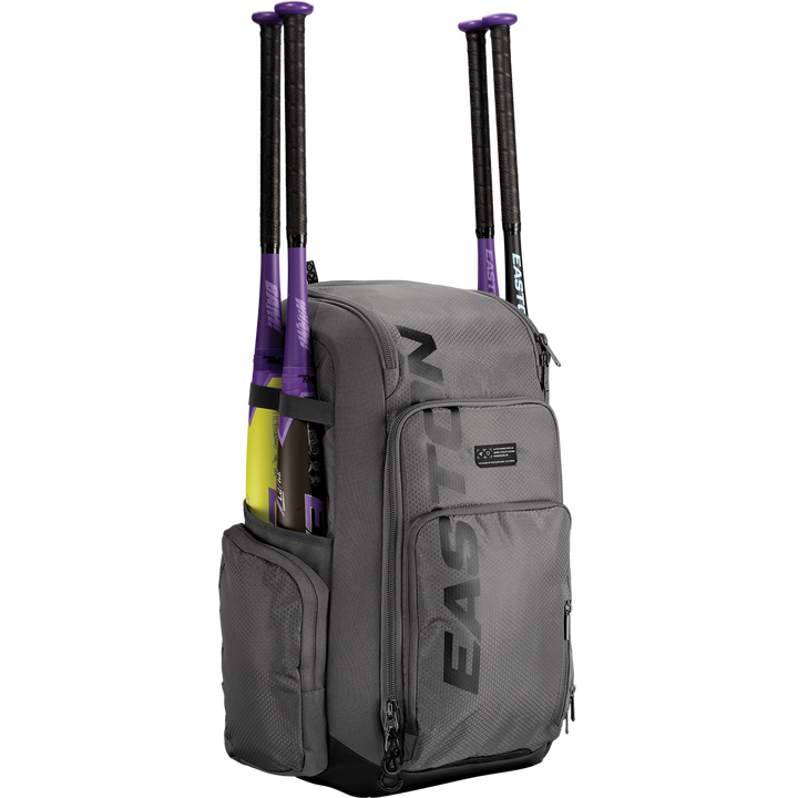 A gray Easton Roadhouse Backpack: EBA004 holds two purple and black baseball bats in side pockets, featuring multiple zippered compartments. The EASTON brand name is printed vertically on the front of this versatile bat bag.