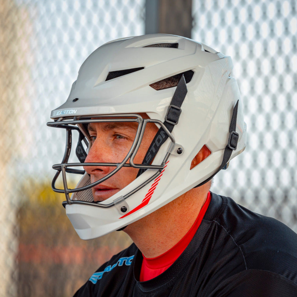 A man wearing an Easton Hellcat Slowpitch Fielding Helmet (EHCATH), featuring a white shell, faceguard, and black chin straps, sits by a chain-link fence. He sports a black shirt with a touch of red at the collar.