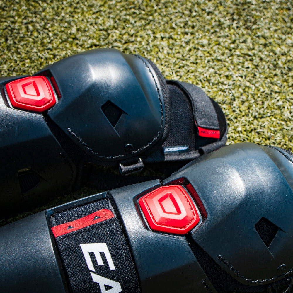 Close-up of black Easton Hellcat Slowpitch Fielding Leg Guards (EHCATL) with red hex accents resting on green turf; the EASTON brand is visible on the strap.
