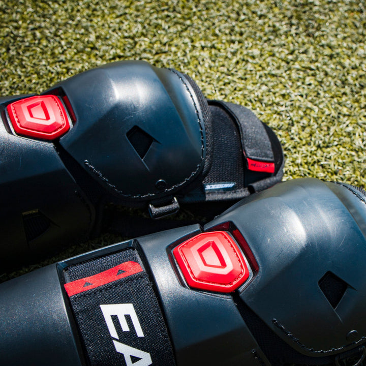 Close-up of black Easton Hellcat Slowpitch Fielding Leg Guards (EHCATL) with red hex accents resting on green turf; the EASTON brand is visible on the strap.