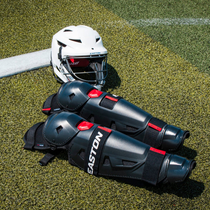 A white helmet and black Easton Hellcat Slowpitch Fielding Leg Guards (EHCATL) by Easton lie on green artificial turf near a white boundary line under bright sunlight.