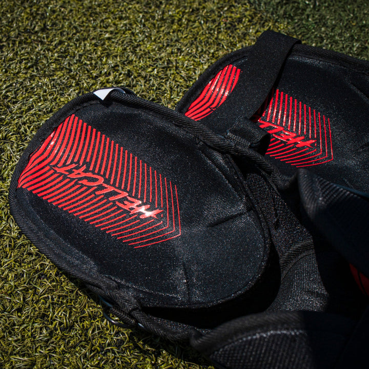 Close-up of Easton Hellcat Slowpitch Fielding Leg Guards (EHCATL) in black with red stripes, featuring Easton branding, lying on green artificial turf.