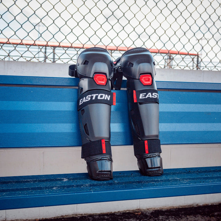 A pair of Easton Hellcat Slowpitch Fielding Leg Guards (EHCATL) in black and red rest upright against blue stadium seats, with a chain-link fence behind—ideal protection for slowpitch softball on game day.