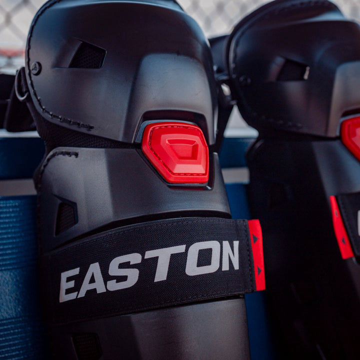 Close-up of black Easton Hellcat Slowpitch Fielding Leg Guards: EHCATL with red accents, displaying the Easton brand on an elastic strap. The leg guards rest on a blue surface near a chain-link fence.
