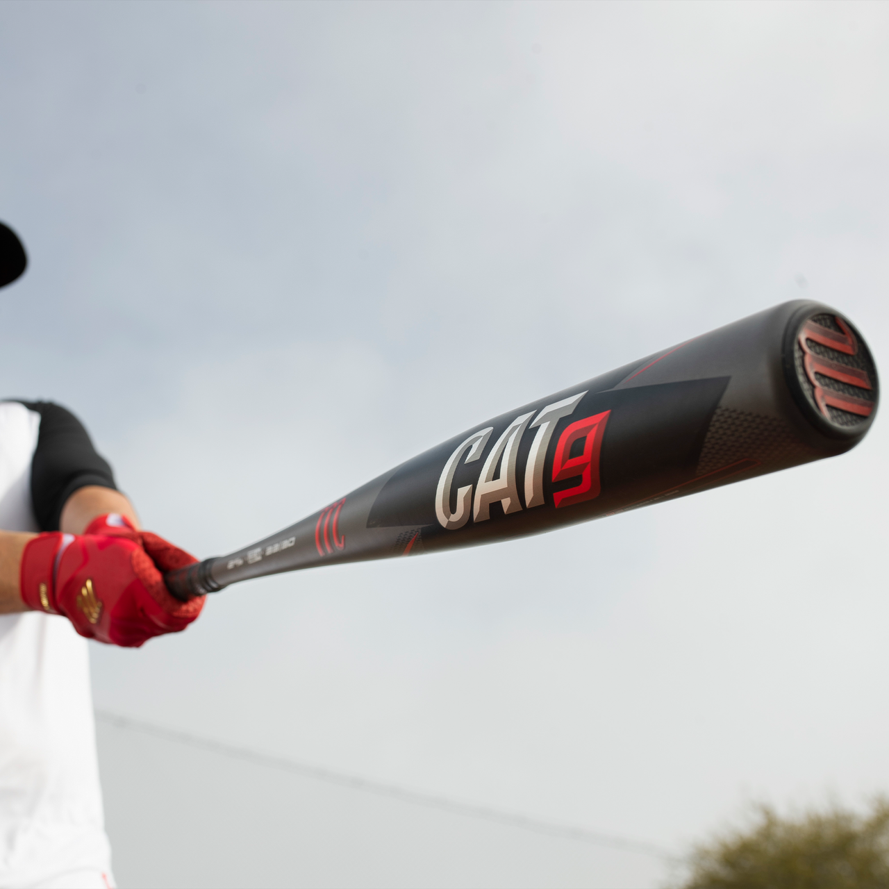 A person in red gloves holds out a black 2021 Marucci CAT9 (-8) 2 3/4" USSSA Baseball Bat (MSBC98, USED) with red and white lettering, by Marucci, against a cloudy sky.
