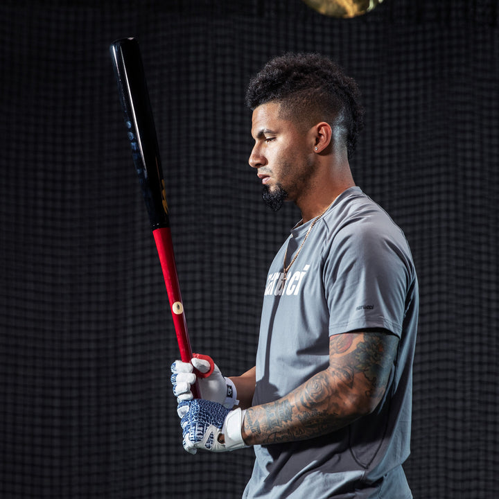 A tattooed baseball player in a gray shirt and gloves holds the Marucci GLEY25 Gleyber Torres Pro Model Maple Wood Bat (MVE2GLEY25-CH/BK) by Marucci, standing in profile against a dark, netted background with a focused look.