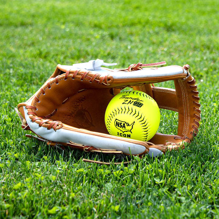 A Dudley NSA Thunder ZN Hard Core ICON 12" 44/400 Composite Slowpitch Softball by Dudley sits inside a brown and white leather glove on green grass outdoors.