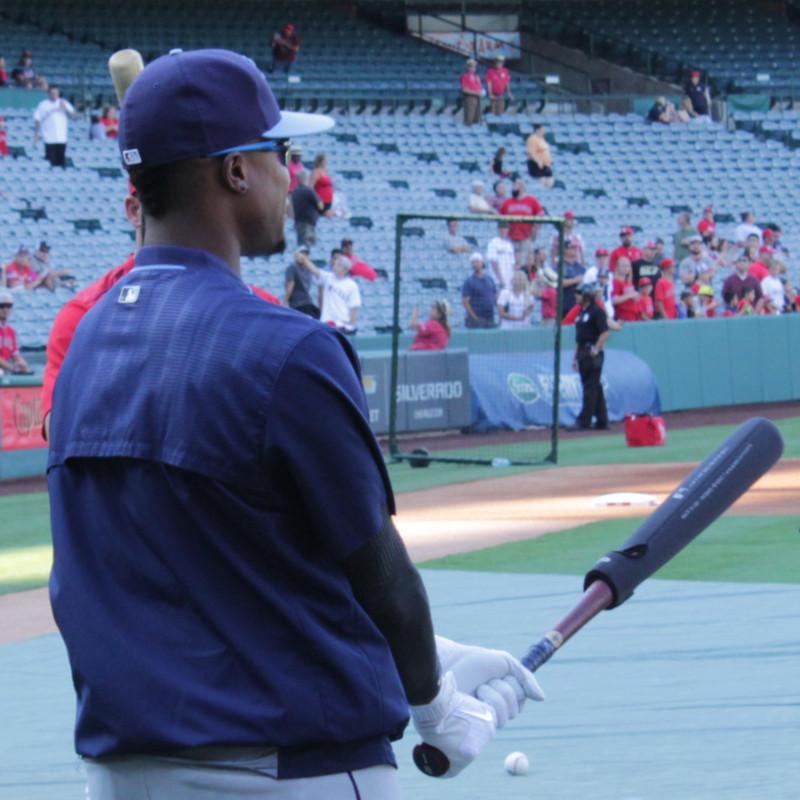 A baseball player in a blue uniform and cap uses the Varo RAP Weighted Hittable Training Sleeve by Varo during batting practice on a field, with empty stadium seats and some fans in the background.