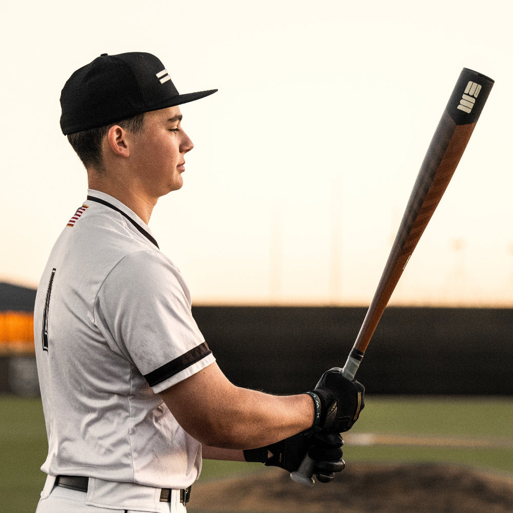 A baseball player in a white uniform and black cap stands on the field gripping a used 2023 Warstic Warhawk3 (-3) BBCOR Baseball Bat: MBWH323WF3, ready to swing. The batâs balanced weight and alloy boost power; the background is out of focus.